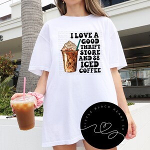 Puede incluir: Camiseta blanca con un gr&aacute;fico de una taza de caf&eacute; helado y el texto "Me encanta una buena tienda de segunda mano y 8 &euro; de caf&eacute; helado". El gr&aacute;fico es en negro y marr&oacute;n. La camiseta la lleva puesta una persona. El texto "Little Black Heart Co." est&aacute; en un c&iacute;rculo negro en la esquina inferior derecha de la imagen.