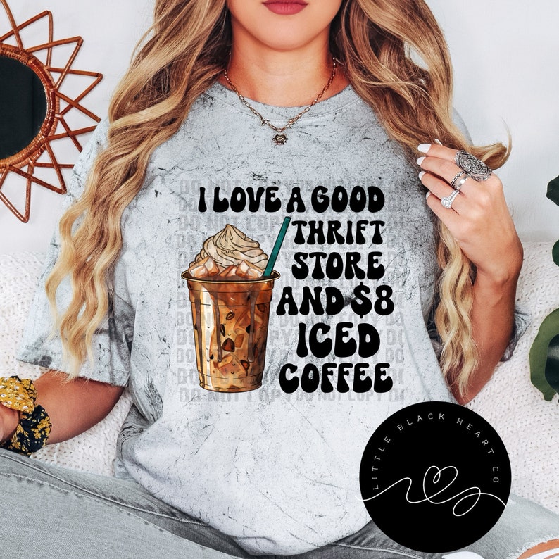 Puede incluir: Una camiseta gris con un gr&aacute;fico de un caf&eacute; helado y el texto "Me encanta una buena tienda de segunda mano y caf&eacute; helado de 8 euros". La camiseta tambi&eacute;n tiene un logotipo circular negro con el texto "Little Black Heart Co."