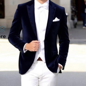 May include: A man wearing a navy blue velvet blazer with a white shirt and white pants. He is wearing a white bow tie and a white pocket square.