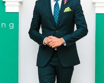 May include: A man wearing a dark green suit with a patterned tie and a green pocket square. He is standing in front of a green wall with the word "Bling" written on it. There is a potted plant with pink flowers to the right.