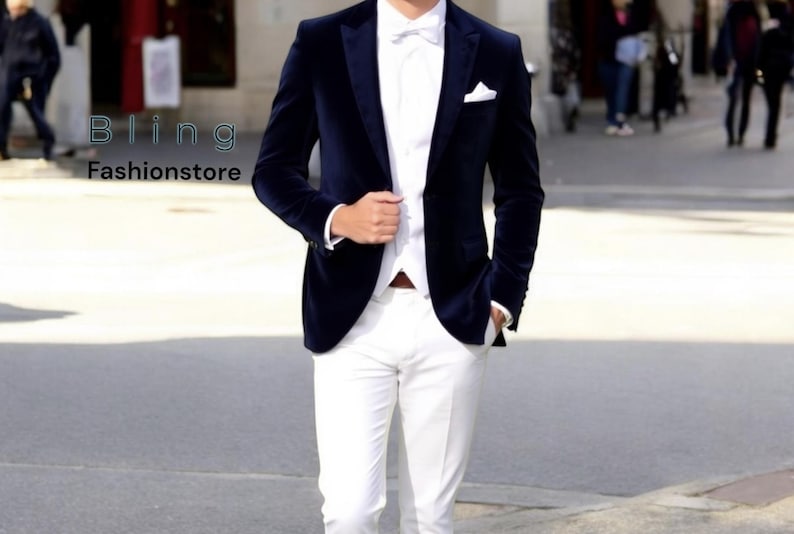 May include: A man wearing a navy blue velvet blazer with a white shirt and white pants. He is wearing a white bow tie and a white pocket square.