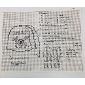 May include: A black and white cross-stitch pattern for a Shriner's Fez. The pattern includes the words "OMAR HEJAZ JAMIL OASIS" and instructions for stitching with DMC floss in various colors.