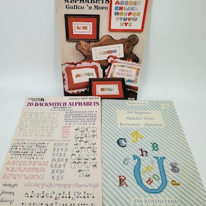 Puede incluir: Se muestran varios libros de patrones de alfabetos de punto de cruz. Las portadas presentan varias fuentes y diseños, incluyendo muestras enmarcadas con palabras como "Love" y "Welcome". Los libros ofrecen una variedad de estilos de alfabeto para proyectos de costura.
