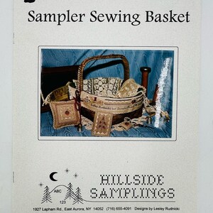 May include: A vintage-style sewing basket with a brown wicker handle and a white fabric lining. The basket is filled with small, embroidered pieces, including a pillow, a small bag, and a needle case. The text "Lesley Rudnicki for Samplings" is visible on the basket.