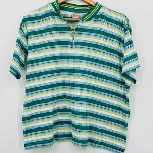 May include: A short-sleeved shirt with a green, white, and blue striped pattern. The shirt has a quarter-zip closure and a ribbed texture. The collar and sleeve edges are trimmed with green and yellow stripes. The shirt is hanging on a wooden hanger.