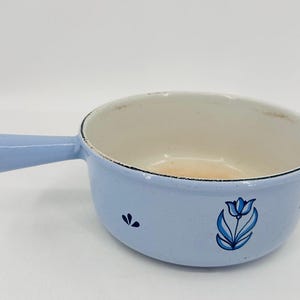 Könnte beinhalten: Ein hellblaues Emaille-Kochgeschirr mit einem einzelnen Griff. Die Pfanne hat einen weißen Innenraum und ist mit einem blauen Tulpenmuster und kleinen floralen Akzenten verziert. Die Pfanne hat einen Durchmesser von etwa 18 cm.