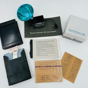 May include: A collection of vintage Polaroid camera accessories, including a flash, a Polaroid Automatic 103 Land Camera manual, a Polaroid Portrait Kit box, and a Cold-Clip instruction card. The items are arranged on a white surface.
