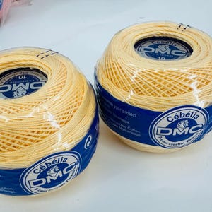 May include: Two balls of pale yellow DMC Cebelia 3-cord crochet thread. Each ball is wrapped in clear plastic and has a blue and white label with the DMC logo and product information. The thread appears to be tightly wound.