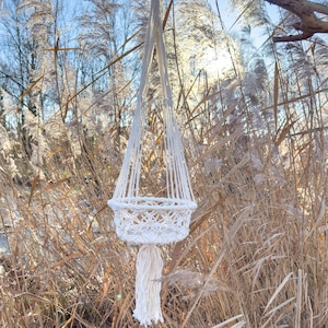 Può includere: Un portapiante in macramè bianco sporco fatto a mano. Il portapiante presenta un design a cestino intrecciato con una lunga nappa sul fondo. Il supporto è sospeso da due cordoncini, perfetto per esporre piccole piante o fiori. Lo sfondo è un ambiente naturale all'aperto.