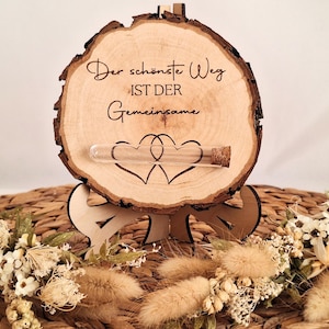 Könnte beinhalten: Ein dekoratives Holzstück mit dem Text "Der schönste Weg ist der Gemeinsame" und zwei ineinandergreifenden Herzen. Eine kleine Glasampulle mit Korken ist befestigt. Das Stück steht auf einem Holzständer und ist von Trockenblumen umgeben.
