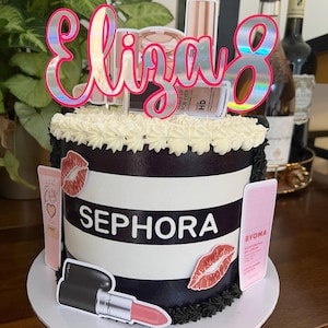 May include: A birthday cake decorated with a Sephora theme. The cake has black and white stripes, with the Sephora logo in the centre. It is adorned with lipstick, makeup, and a topper that says "Eliza 8". The cake is on a white plate.