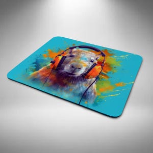 May include: A square mouse mat featuring a colourful illustration of a capybara wearing orange headphones. The capybara is set against a bright blue background with paint splatter effects. Designed for computer use.