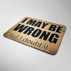 May include: A rectangular mouse mat with a wood-grain effect. The text "I MAY BE WRONG but I doubt it" is printed in bold black. The mat has rounded corners and a white border, a practical desk accessory.
