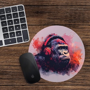 May include: Circular mouse mat with a gorilla wearing red headphones. The gorilla is illustrated in a vibrant, artistic style with red, purple, and orange tones. A black mouse and keyboard are on a wooden surface.