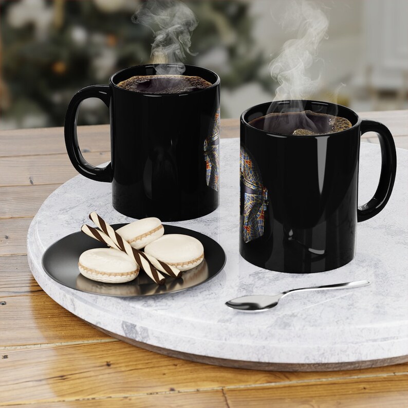 May include: Two black coffee mugs with a colorful design on the side. The mugs are filled with coffee and steam is rising from both. The mugs are on a white marble surface with a small plate of cookies and a spoon.