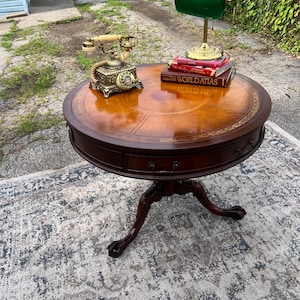May include: A round wooden drum table with a leather top, featuring a drawer with metal hardware and a tripod base with claw feet. On the table sits a vintage-style rotary telephone, a green banker lamp, and a World Atlas book.