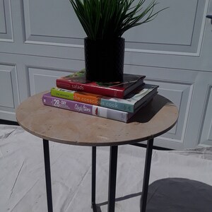 May include: A round, light brown table with a black metal base. Three cookbooks are stacked on the table, with the top book titled "salads". A potted green plant sits on top of the books.