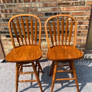 May include: Two wooden bar stools with a brown stain finish. The stools have a slatted back and a round seat. The stools are sitting on a gray surface.