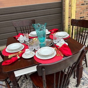 May include: A wooden table set with four chairs, white and red placemats, white plates with gold trim, red napkins, clear glass water goblets, and a blue glass vase.