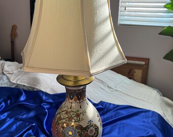 May include: A white ceramic table lamp with a gold and floral design. The lamp has a white fabric lampshade with a gold trim. The lamp is sitting on a blue satin fabric.