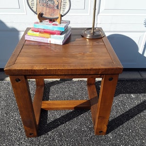 May include: A wooden end table with a brown finish. The table has a square top and a simple, open frame. There are four books and a decorative plate on top of the table.