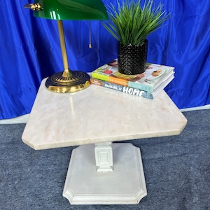 May include: A white marble end table with a white painted base. A green glass lamp with a brass base sits on the table, along with a stack of books. A potted plant with green leaves sits on the table.