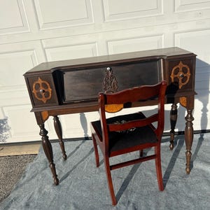 May include: A dark brown antique desk with a matching chair. The desk has decorative wood accents and a carved center panel. The chair has a dark seat and a curved back. The desk and chair are set against a white garage door.