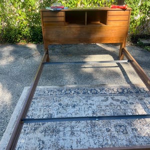 May include: A wooden bed frame featuring an integrated headboard with built-in storage compartments. The headboard includes two side sections with horizontal slats and a central open shelving area. The frame is constructed from brown wood.