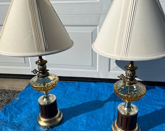 May include: A pair of vintage table lamps with amber glass bases and white drum shades. The lamps have a gold metal finish and a wooden base.