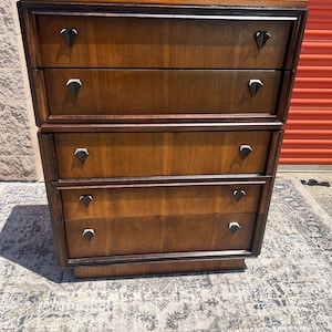May include: A tall wooden dresser featuring five horizontal drawers with a dark brown finish. Each drawer is equipped with two distinct triangular-shaped black metal pulls. The piece has a flat top surface and a recessed base, showcasing a wood grain.