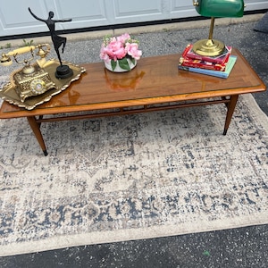 May include: A rectangular wooden coffee table with a glass top, tapered legs, and a lower shelf. On the table are a gold-toned rotary telephone on a tray, a black dancer figurine, a white bowl with pink flowers, and a green banker lamp on books.