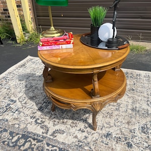 May include: A two-tiered, round wooden side table with a glossy finish. A green glass desk lamp, books, and decorative items sit on the top tier. The table is on a patterned rug.