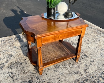 May include: A wooden side table with a glossy finish, featuring a lower shelf with a woven cane insert. A silver tray with decorative items sits on top. The table has a drawer and is set on a patterned rug.