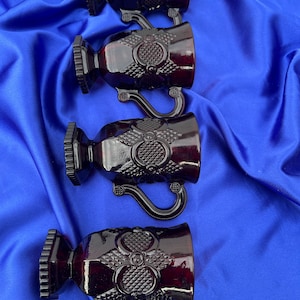 May include: Four dark red goblets with ornate detailing. Each goblet features a decorative handle, a textured base, and a detailed design on the cup. The goblets are arranged on a blue fabric background.