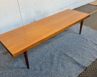 May include: A mid-century modern coffee table with a rectangular light brown wooden top and four tapered dark brown legs. The table is approximately 60 inches long.