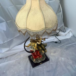 May include: A decorative table lamp with a cream-colored lampshade featuring a floral pattern and beaded fringe. The base depicts two figures playing musical instruments. The base is mounted on a dark, rectangular platform.