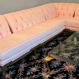 May include: A large L-shaped sectional sofa upholstered in light pink fabric with a tufted backrest and a dark wood frame. Beside it sits a rectangular coffee table featuring a clear glass top and a gold-toned metal base with curved legs.