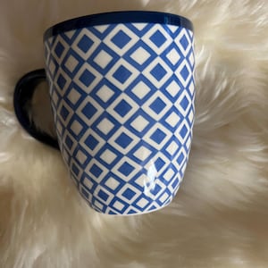 May include: A white ceramic mug with a blue geometric pattern. The mug has a blue rim and a blue handle.