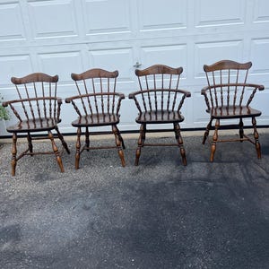 May include: Four wooden Windsor-style chairs with a dark brown finish. Each chair features a curved backrest with spindles, armrests, and turned legs. The chairs are arranged in a row, showcasing their classic design.