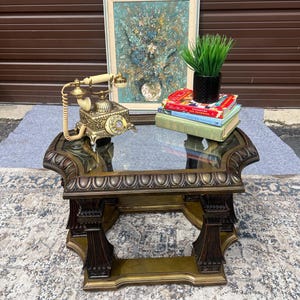 May include: A square end table with a glass top and a dark bronze-toned ornate base. On the table sits a vintage-style rotary telephone with a cream handset, a black pot containing green grass, and a stack of three hardcover books.