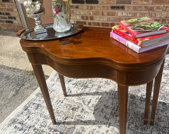 May include: A dark wood side table with a curved top and three legs. The table has a decorative inlay around the edge. A silver tray with a floral pitcher and a vase with white flowers is on the table. A stack of cookbooks is on the table.