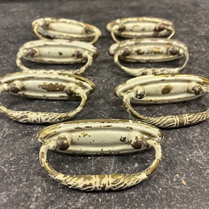 May include: Eight antique white and gold drawer pulls with a decorative design. The pulls are oval shaped with two knobs on each side.