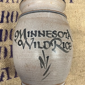 May include: A white ceramic jar with a black rim and a blue stripe. The jar has a handwritten inscription that reads "Minnesota Wild Rice".