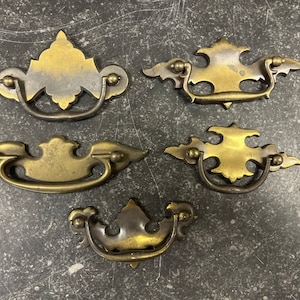 May include: Five antique brass cabinet pulls with ornate designs. Each pull has a curved handle and decorative wings.