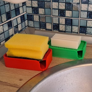 Könnte beinhalten: Eine Küchenszene mit einem roten Schwammhalter mit gelben Schwämmen, einem grünen Halter mit einem weißen Seifenstück und einem Edelstahlbecken. Der Hintergrund zeigt blau-weiß gemusterte Fliesen.