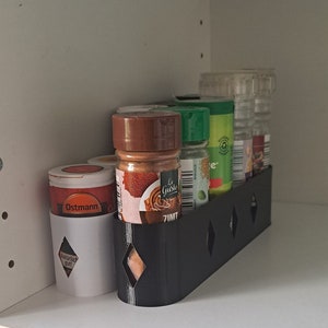 Könnte beinhalten: Ein schwarzer Gewürzregal mit sechs Glasgewürzgläsern. Die Gläser sind mit verschiedenen Gewürzen gefüllt und haben Etiketten mit Text in verschiedenen Sprachen. Das Regal ist an einem weißen Regal befestigt.