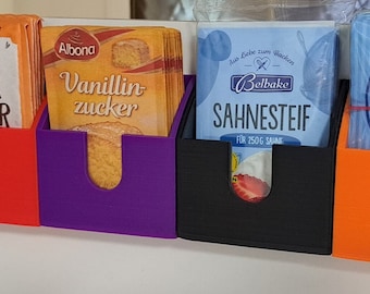 Op de afbeelding: Zes kleine, kleurrijke, aan de muur gemonteerde opbergcontainers van plastic met verschillende bakproducten en een blauwe doos met heldere cakemix. De containers zijn gelabeld met "Hefe", "Back Pulver", "Vanillin Zucker", "Sahnesteif" en "Tortenguß klar".