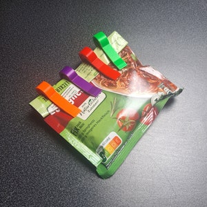 Könnte beinhalten: Ein Set aus roten, grünen, violetten und orangenen Plastik-Beutelclips hält eine Packung Spaghettisoße. Die Packung hat einen grünen Hintergrund mit einem Bild von Spaghetti und Tomaten. Der Text auf der Packung lautet "LECKER FIX noch dazu: mit Natürlichen Zutaten 250 g Spaghetti 200 g mageres Hackfleisch NUTRI-SCORE BCDE".