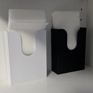Könnte beinhalten: Zwei Taschentuchbox-Halter, einer weiß und einer schwarz, jeweils mit einer gebogenen Öffnung. Beide Halter sind mit weißen Taschentüchern gefüllt. Die Halter sind rechteckig und stehen auf einer weißen Oberfläche.