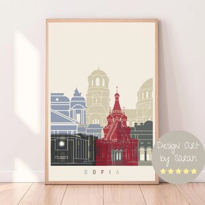 Puede incluir: Una ilustración minimalista de la ciudad de Sofía, Bulgaria, con varios edificios en tonos azules, rojos y grises sobre un fondo beige. El texto "SOFIA" está impreso debajo de la ilustración.